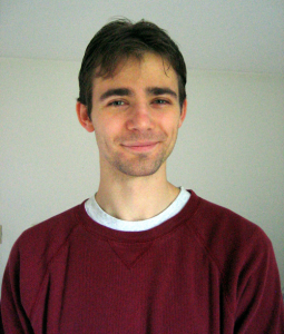 A young man with short brown hair, wearing a maroon sweatshirt over a white shirt, is standing indoors and smiling slightly at the camera.