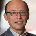 Man with glasses, short receding hair, and a slight smile wearing a suit jacket and collared shirt, posing in front of a blurred background.