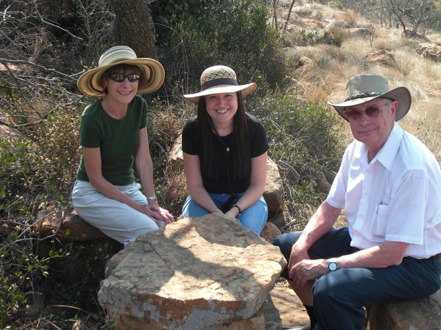 Three people wearing sun hats sit outdoors on rocks, smiling at the camera. Dry grass, bushes, and trees surround them in a sunny, natural setting.