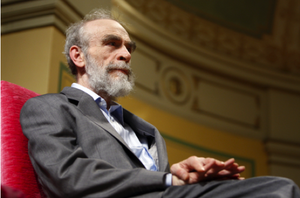 An older man with a gray beard and suit sits on a red chair, looking thoughtful. The background features ornate architectural details and warm lighting.