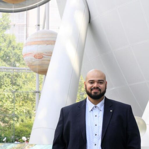 A man in a blazer stands indoors, smiling, with large models of planets and a white spherical structure behind him.