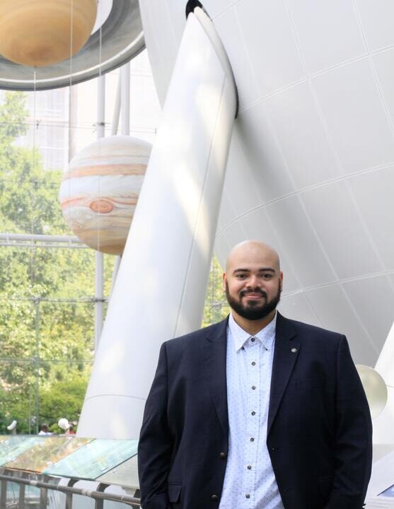 A man in a blazer stands indoors, smiling, with large models of planets and a white spherical structure behind him.