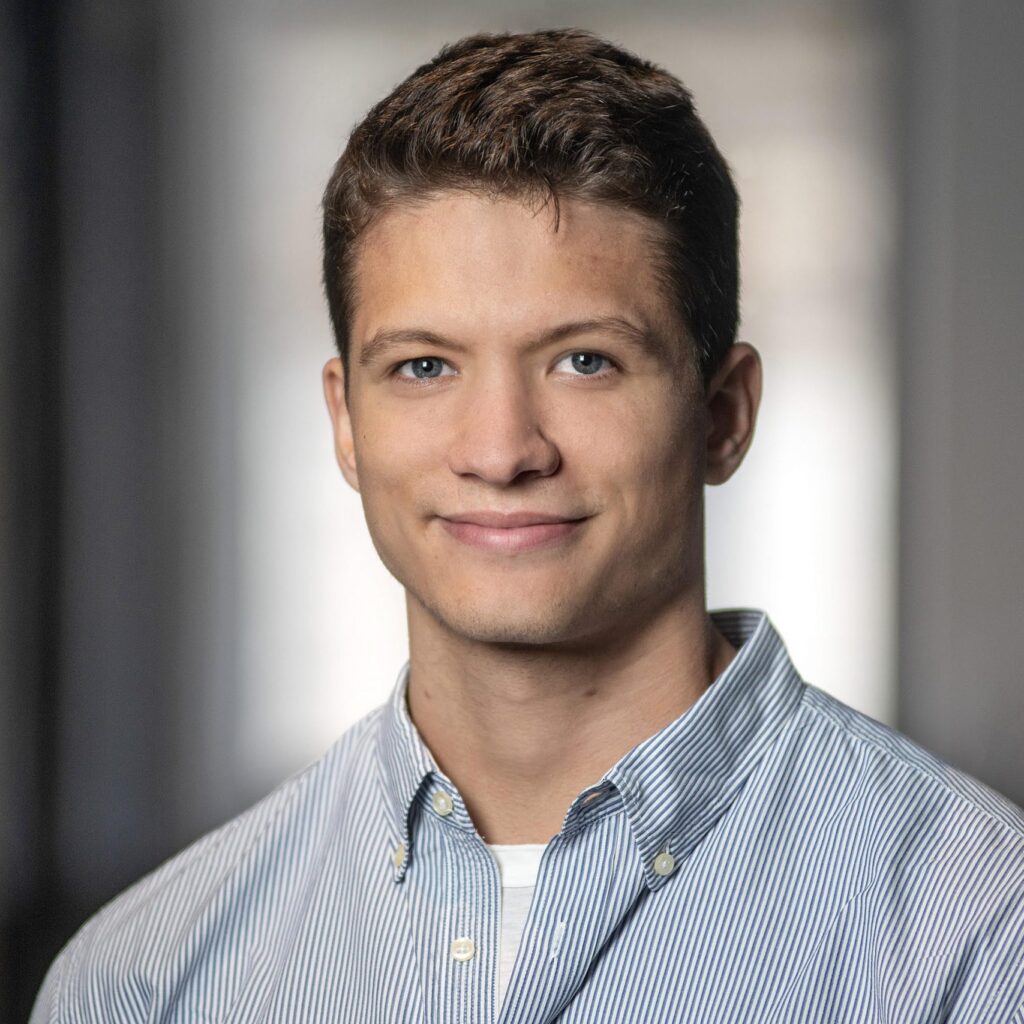 A young man with short brown hair, wearing a light blue striped button-down shirt, smiles softly while standing indoors with a blurred background.