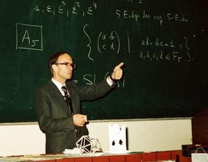 A man in a suit stands in front of a chalkboard filled with mathematical equations, giving a thumbs-up. Geometric models sit on the table beside him.