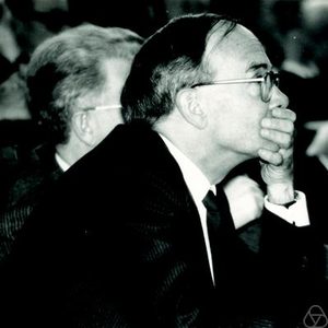 Black-and-white photo of a man in a suit and glasses, sitting with his hand over his mouth, appearing deep in thought. Another man is partially visible in the background.