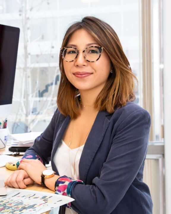 A woman with shoulder-length brown hair and glasses sits at a desk in an office, smiling at the camera. She wears a blazer and holds a colorful sheet of paper. A computer and books are on the desk.