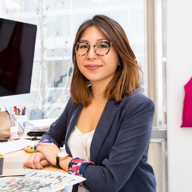 A woman with shoulder-length brown hair and glasses sits at a desk in an office, smiling at the camera. She wears a blazer and holds a colorful sheet of paper. A computer and books are on the desk.