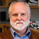An older man with a white beard, wearing a brown jacket over a striped shirt, stands indoors with shelves and books in the background.