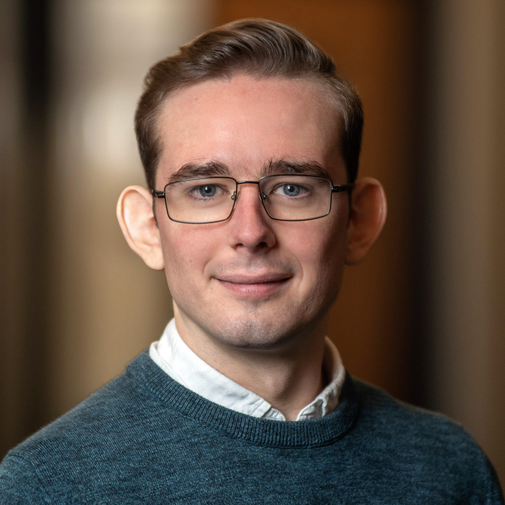 A young man with short light brown hair, glasses, and a teal sweater over a white collared shirt smiles softly at the camera. The background is softly blurred.