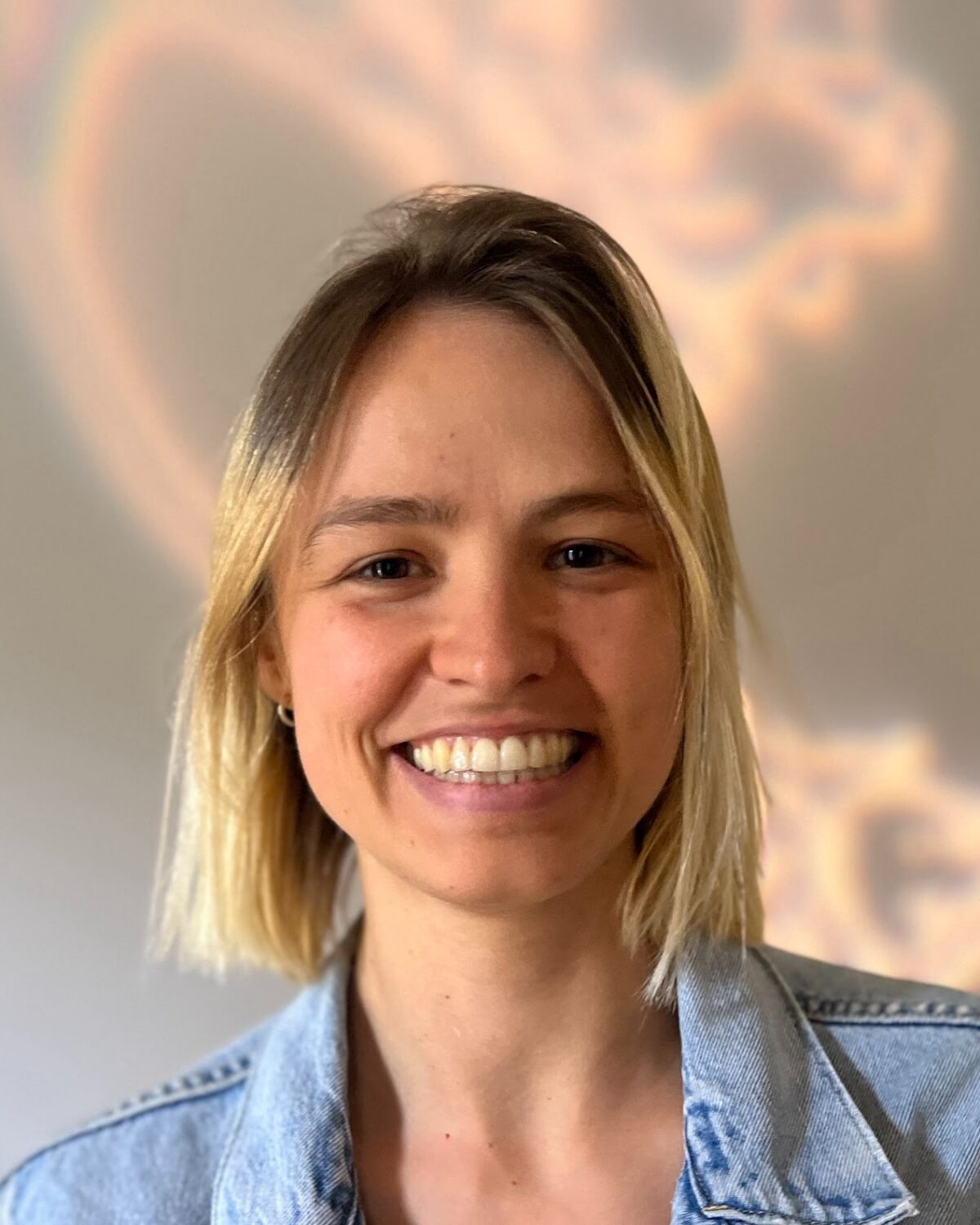 A person with straight, shoulder-length blonde hair smiles warmly. They are wearing a light denim jacket. The background is softly blurred with a swirl of light and pastel colors.