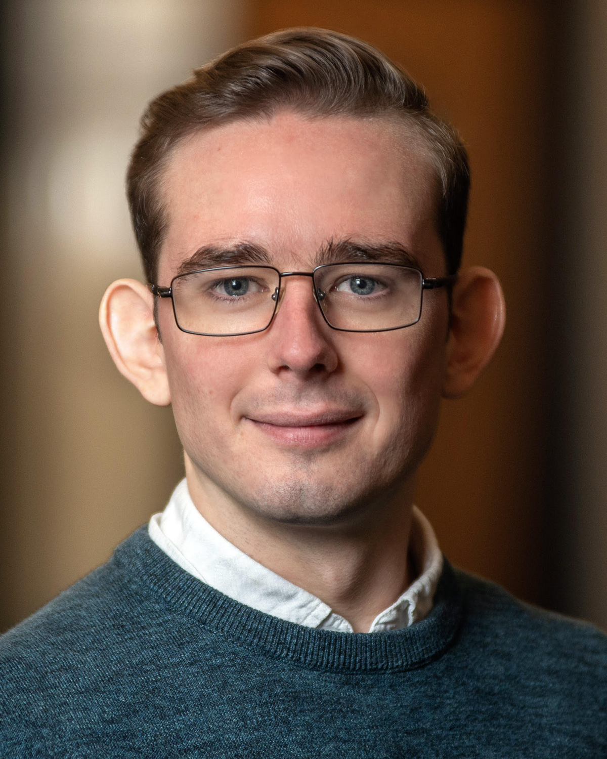 A young man with short light brown hair, glasses, and a teal sweater over a white collared shirt smiles softly at the camera. The background is softly blurred.