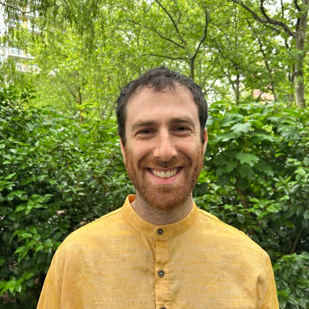 A man wearing a yellow shirt stands outside in front of green bushes and trees, smiling at the camera.