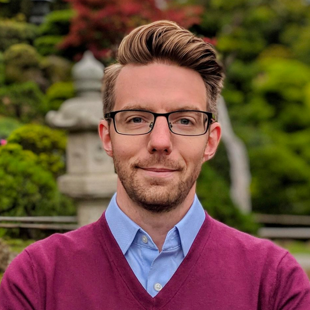 A man with glasses and short, styled hair wears a light blue shirt and maroon sweater. He stands outdoors with a soft-focus garden background, including greenery and a stone lantern.