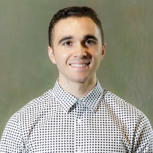 A person with short dark hair smiles at the camera, wearing a white button-up shirt with a black geometric pattern, standing in front of a plain, greenish background.