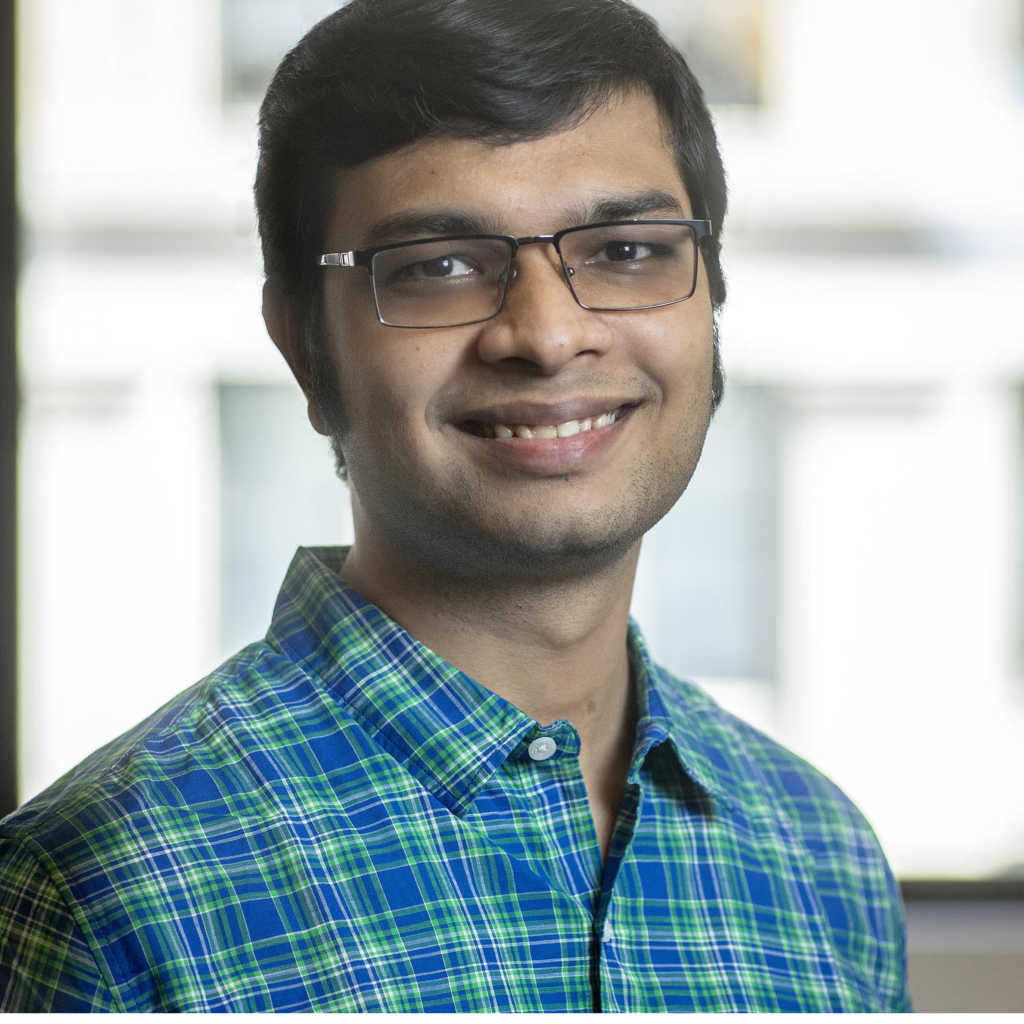 A man with short dark hair and glasses, wearing a blue and green plaid shirt, smiles at the camera. The background is softly blurred, showing large windows with bright daylight.