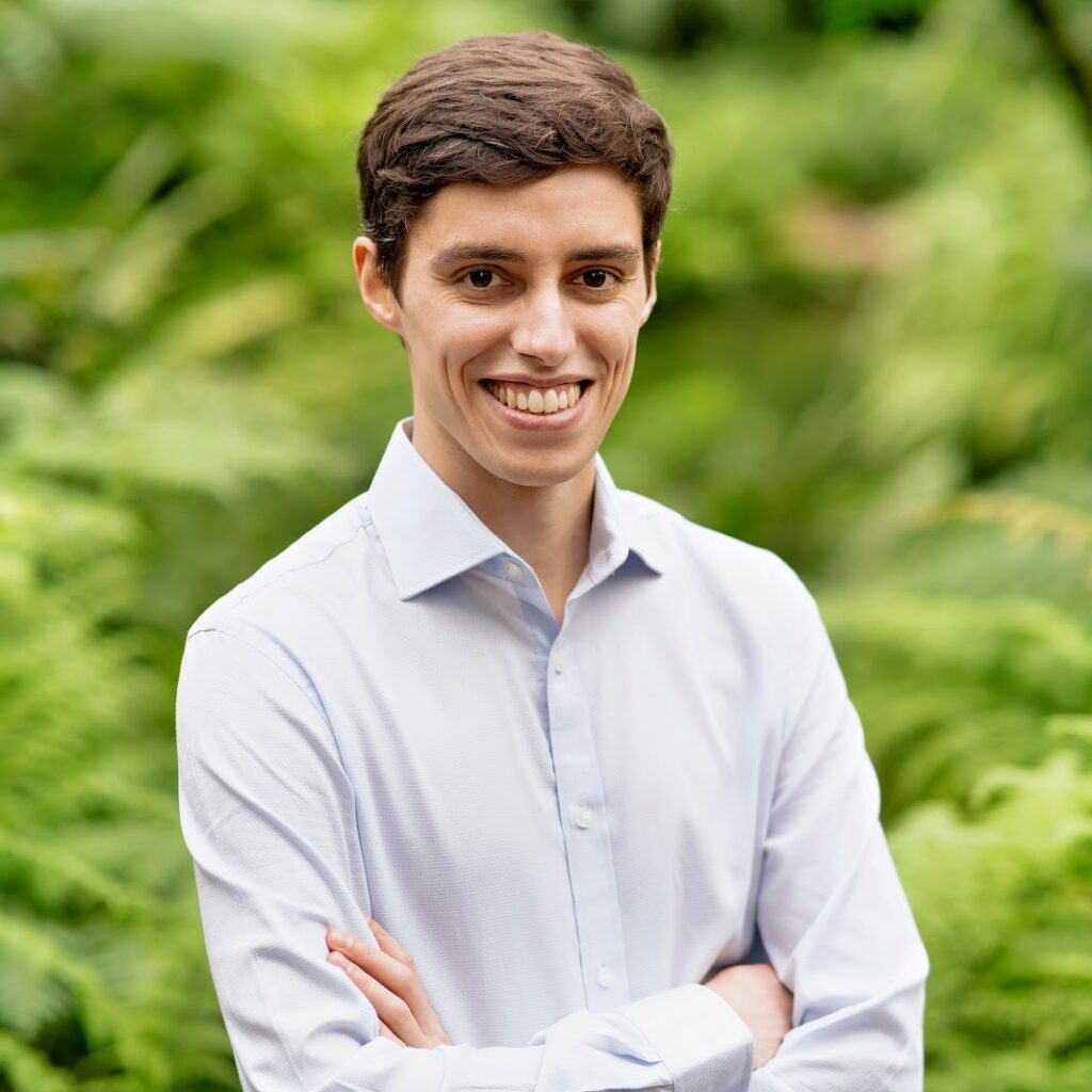A young man with short brown hair, wearing a light blue dress shirt, stands outdoors with arms crossed and smiles, with lush green foliage in the background.
