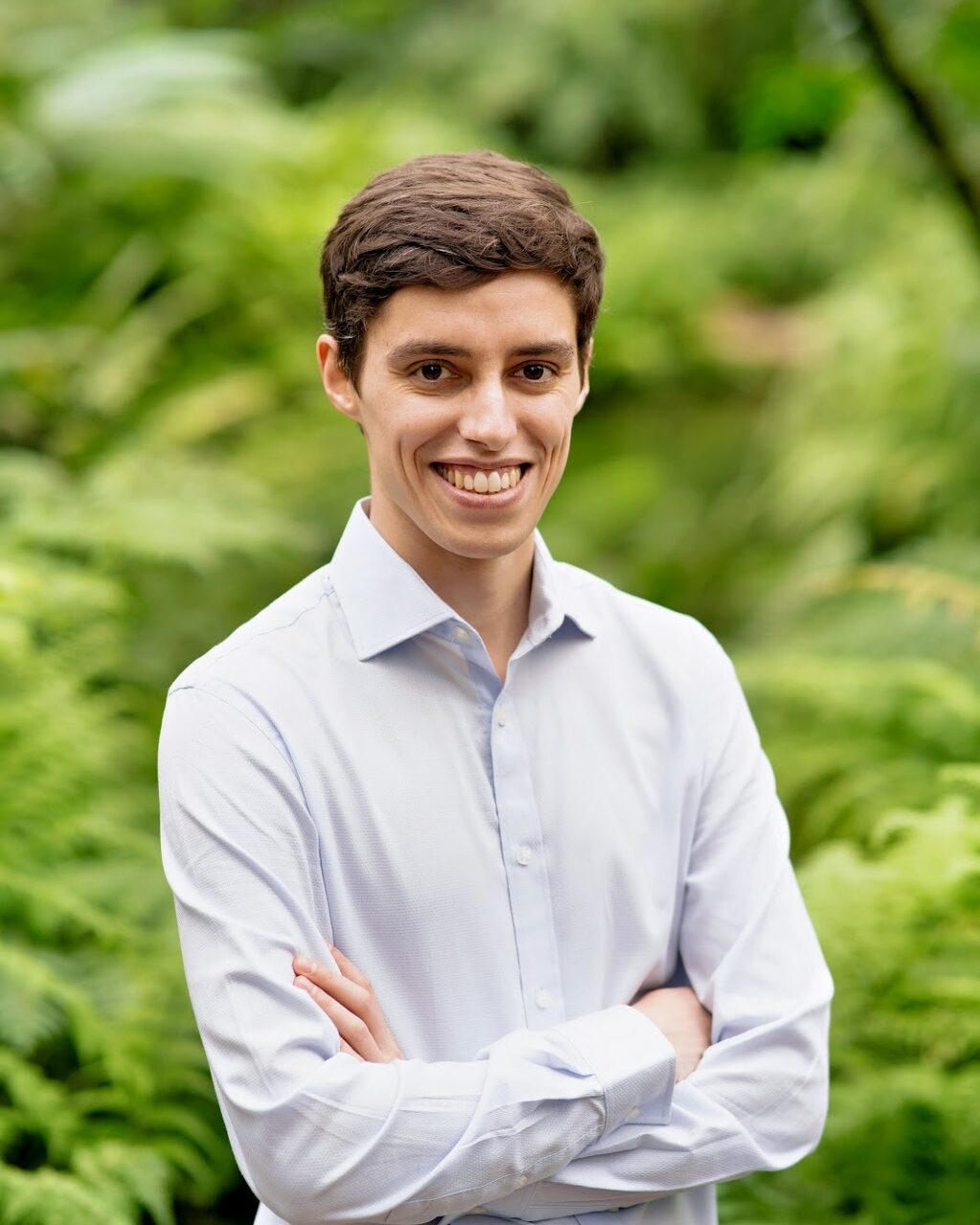 A young man with short brown hair, wearing a light blue dress shirt, stands outdoors with arms crossed and smiles, with lush green foliage in the background.