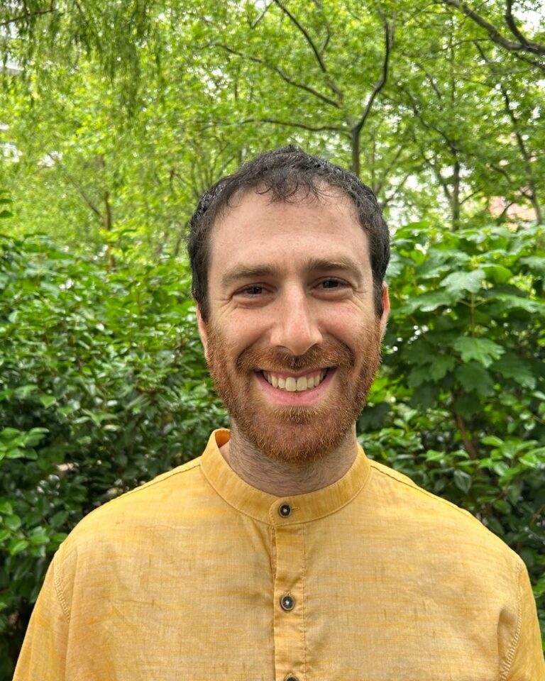 A man wearing a yellow shirt stands outside in front of green bushes and trees, smiling at the camera.