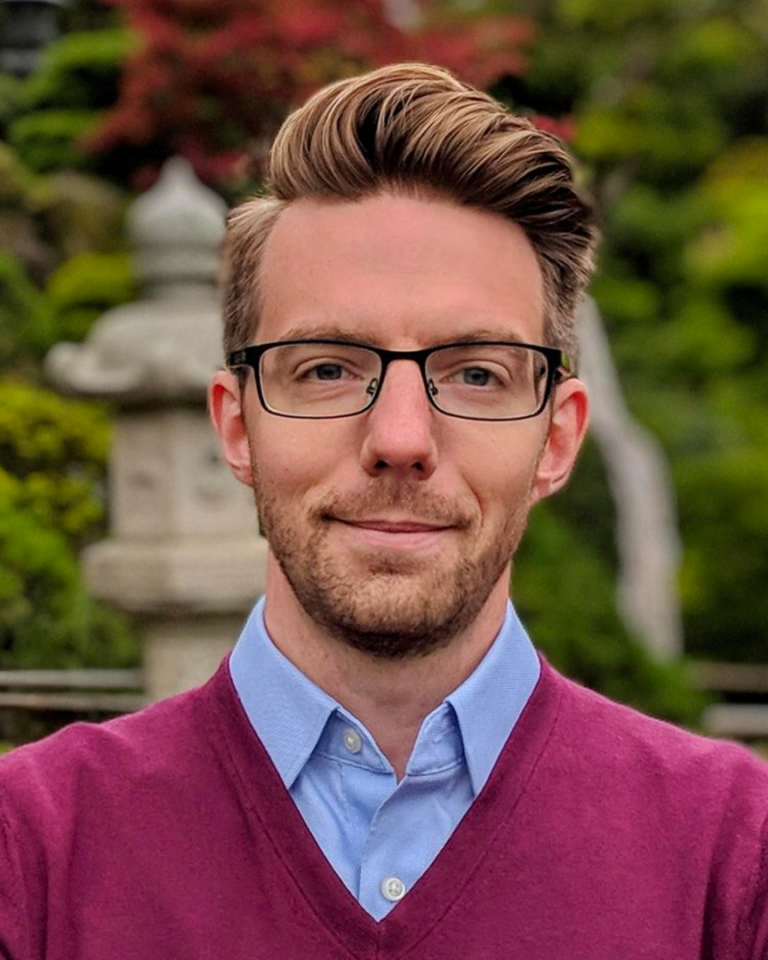 A man with glasses and short, styled hair wears a light blue shirt and maroon sweater. He stands outdoors with a soft-focus garden background, including greenery and a stone lantern.