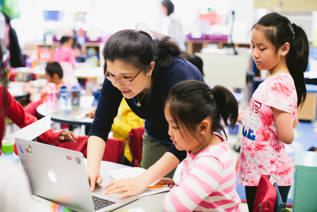A teacher leans over to a laptop as two children stand next to her