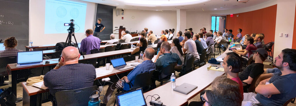 A group of people sit in a classroom, watching a presenter speak next to a projection screen with diagrams. Laptops are open on desks, and a camera records the session.