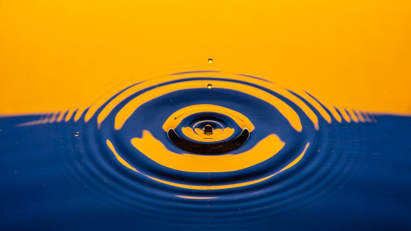 A photograph of what appears to be a drop of water breaking the surface of a pool of water and causing concentric circular ripples to form around the site of the impact.