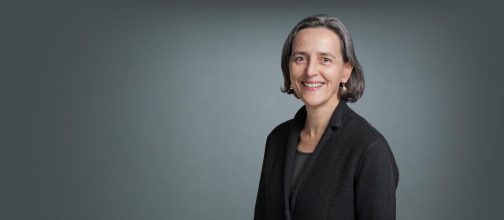 A woman with short gray hair, wearing a black blazer and dark top, stands smiling against a plain gray background.