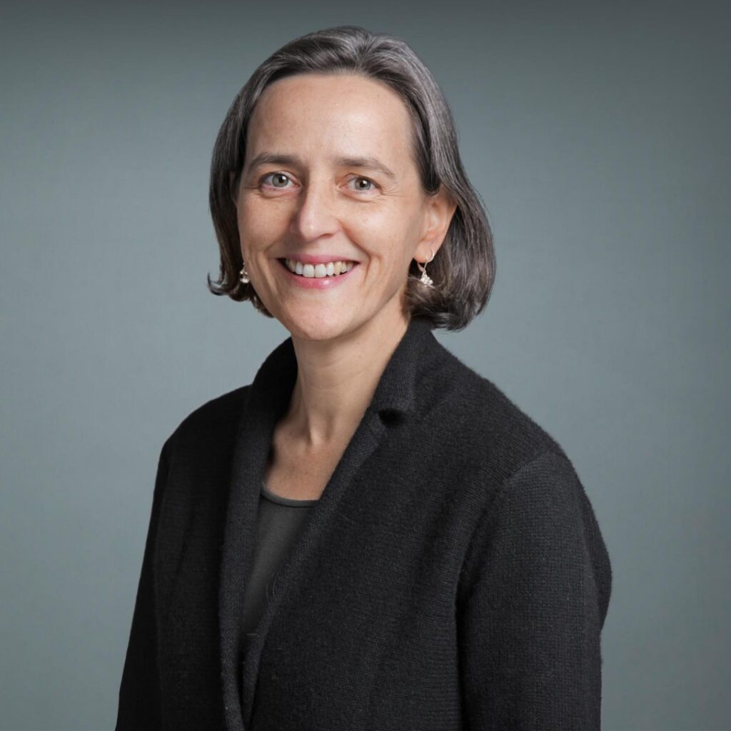A woman with short brown hair and a black blazer smiles at the camera. The background is plain and grey.