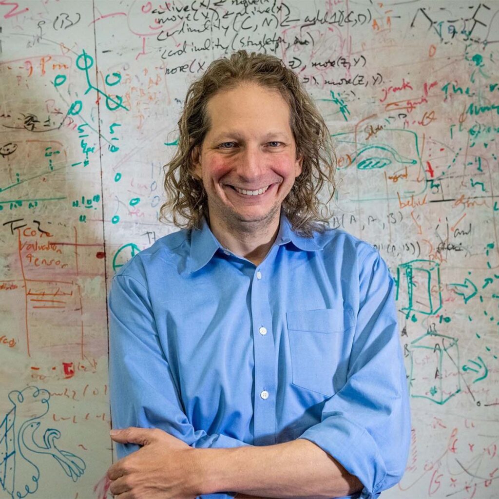 A photograph of Joshua Tenenbaum portrait in front of a whiteboard with various equations, shapes, and other symbols written on it with marker.A photograph of Joshua Tenenbaum portrait in front of a whiteboard with various equations, shapes, and other symbols written on it with marker.