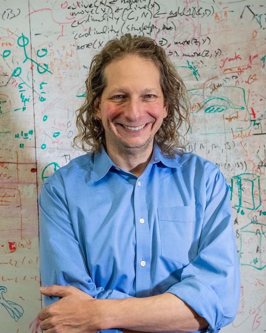 A photograph of Joshua Tenenbaum portrait in front of a whiteboard with various equations, shapes, and other symbols written on it with marker.A photograph of Joshua Tenenbaum portrait in front of a whiteboard with various equations, shapes, and other symbols written on it with marker.