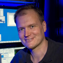 A person with short light brown hair smiles in front of monitors displaying blue and white screens in a dark room.