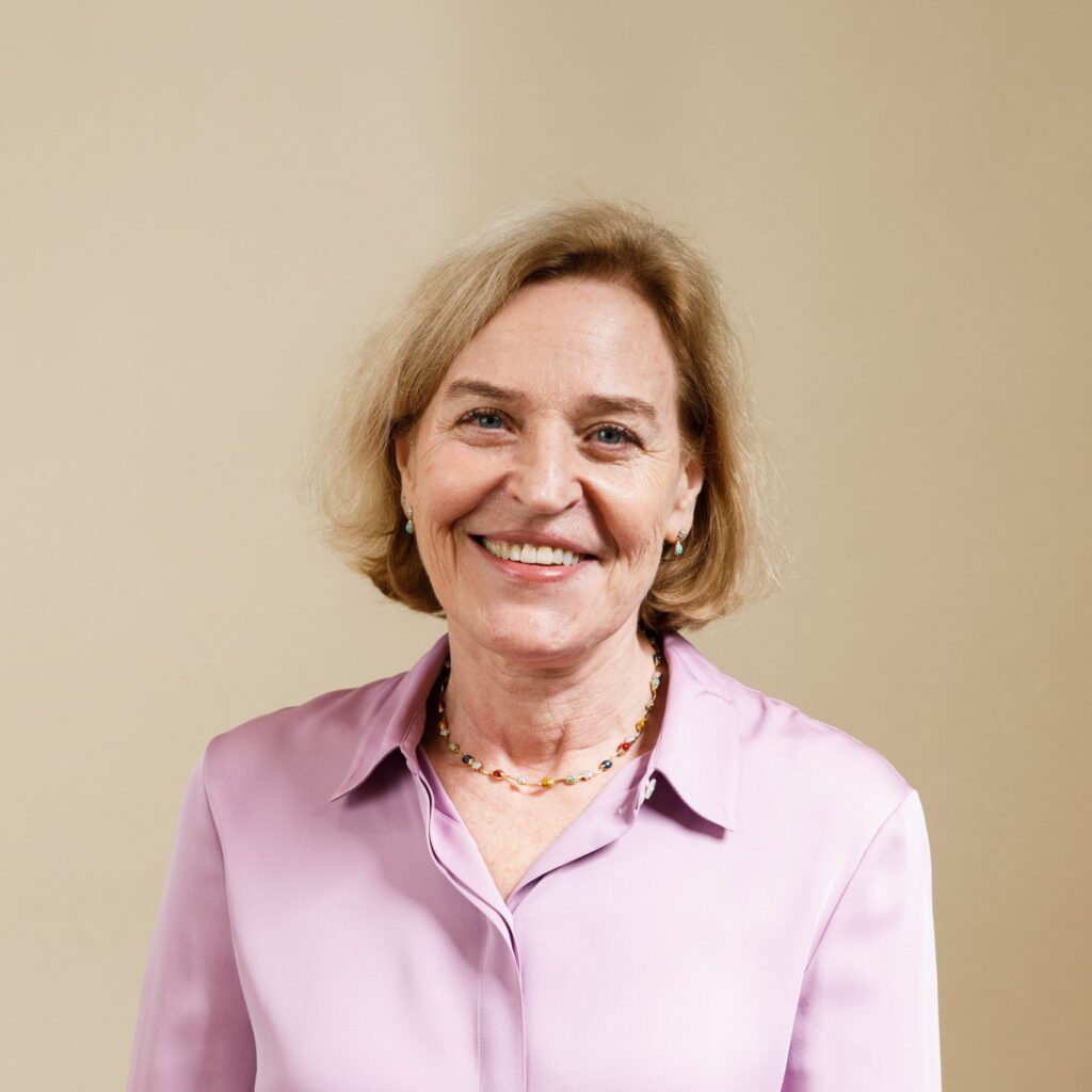 Smiling older woman with short blonde hair wearing a light pink blouse and a colorful beaded necklace, posed against a plain beige background.