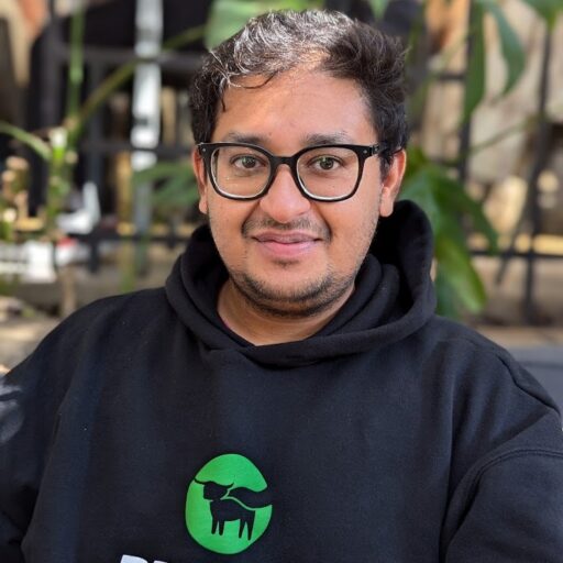 A person with glasses and short dark hair wears a black Beyond Meat hoodie, sitting outdoors with green plants and blurred background visible behind them.