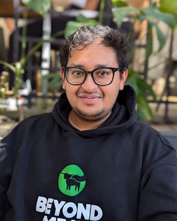A person with glasses and short dark hair wears a black Beyond Meat hoodie, sitting outdoors with green plants and blurred background visible behind them.