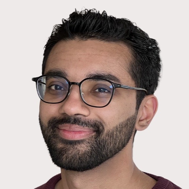 A person with short dark hair, a trimmed beard, and black-rimmed glasses smiles softly at the camera against a plain light background.