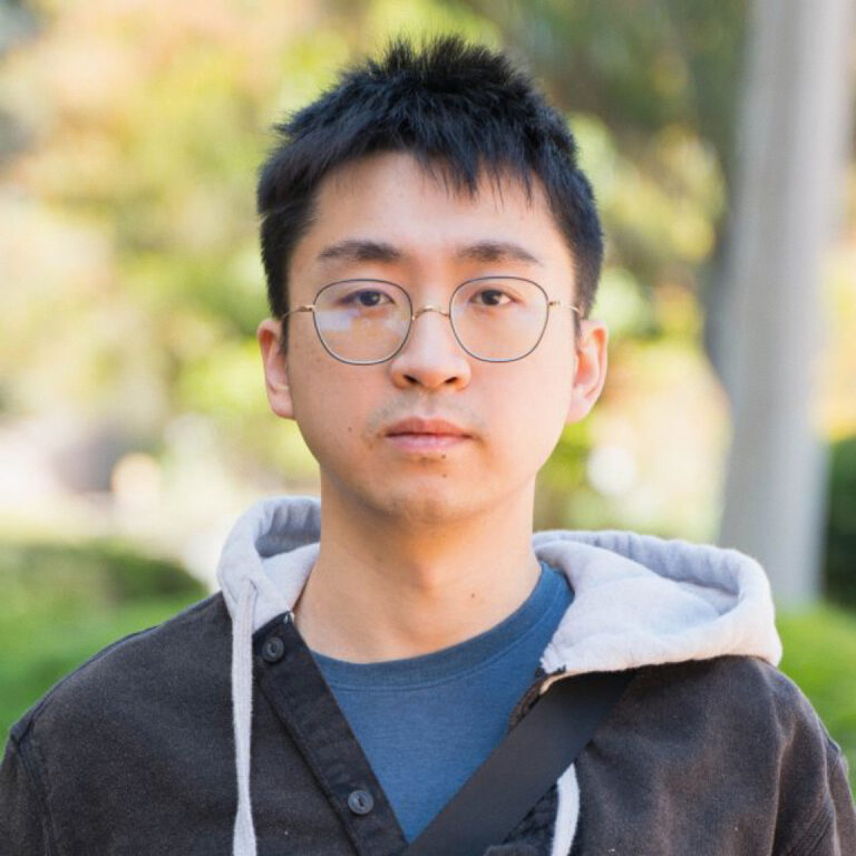 A young person with short black hair, wearing round glasses, a blue shirt, and a dark jacket with a gray hood, stands outdoors with greenery and trees blurred in the background.