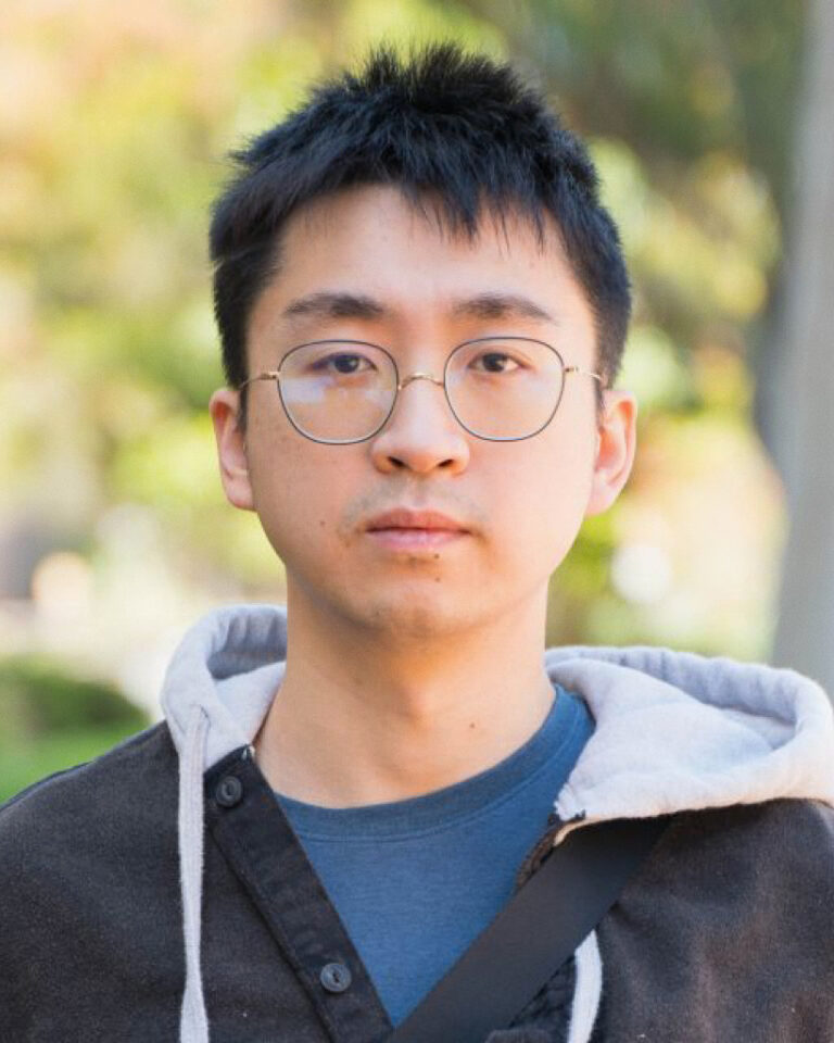 A young person with short black hair, wearing round glasses, a blue shirt, and a dark jacket with a gray hood, stands outdoors with greenery and trees blurred in the background.