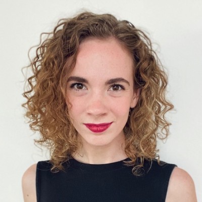 Woman with curly, shoulder-length hair and red lipstick, wearing a sleeveless black top, standing against a plain white background, looking directly at the camera with a slight smile.