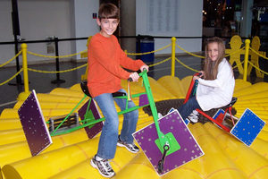 Two children are riding pedal-powered seesaws with square wheels on a yellow, bumpy platform in an indoor play area.