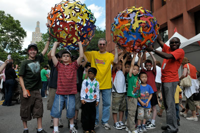 A group of children and adults stand outdoors, holding up two large, colorful sculptures made of interlocking human figures. People smile, and trees and a red building are in the background.