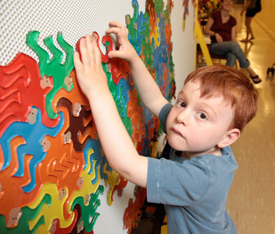 A young boy touches colorful, interlocking animal-shaped pieces on a wall-mounted puzzle, while people are visible in the background.