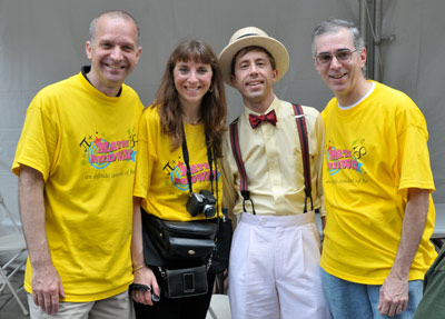 Four adults pose together, three wearing bright yellow event T-shirts and one in a light yellow shirt, white pants, suspenders, and a straw hat with a red bow tie. They are smiling in an outdoor setting.