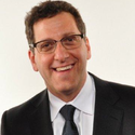 A smiling man with short dark hair and glasses wears a dark suit, white shirt, and dark tie against a plain white background.