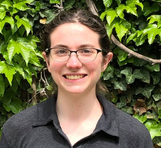 A young person with glasses and brown hair stands outside, smiling, in front of a background of green ivy leaves.