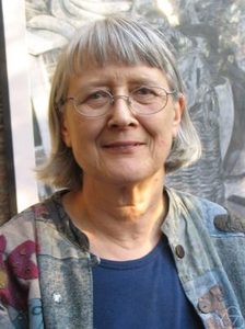 An older person with short gray hair, glasses, and a patterned jacket over a blue shirt is smiling gently in front of a grayscale background.
