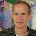 A middle-aged man with short light brown hair, wearing a dark patterned shirt, stands in front of a colorful abstract painting.
