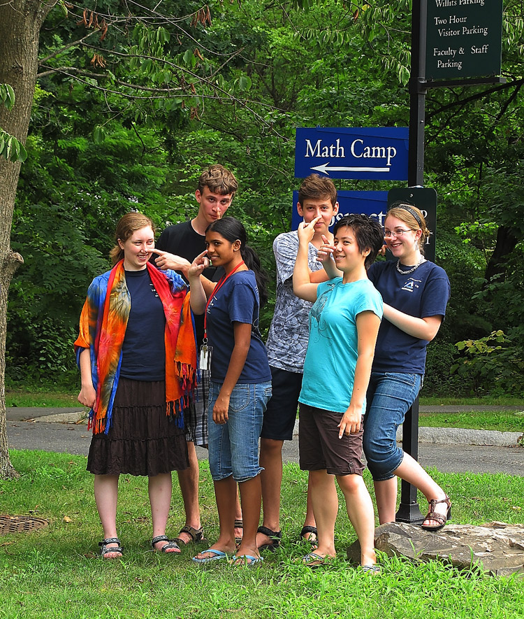 Six teens stand and smile near a Math Camp sign outdoors, with trees and grass in the background. Some are pointing and laughing, while others casually pose, creating a fun and friendly group atmosphere.