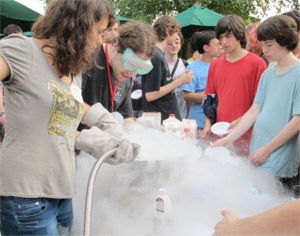 A group of teens gathers around a table with fog or vapor spilling out, while one person wears goggles and gloves, possibly doing a science experiment outdoors.