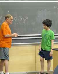 An older man in an orange shirt talks to a boy in a green shirt in front of a chalkboard with drawings and equations, inside a classroom.