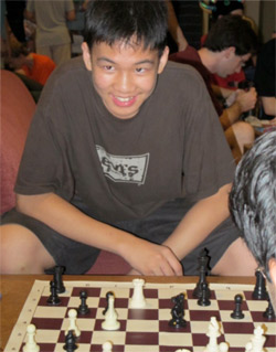 A smiling boy sits at a chessboard, wearing a brown t-shirt and shorts, with chess pieces laid out mid-game. Other people are blurred in the background.